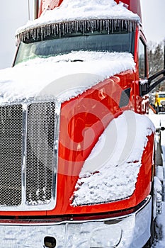 Red big rig semi truck with bright chrome grille covered with snow and icicles standing on the parking lot waiting for the better