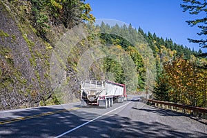 Red big rig semi truck with bulk semi trailer running downhill on the winding autumn road with rock mountain wall and forest