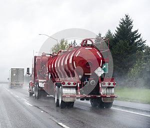 Red big rig semi Truck with tank semi trailer for transportation of chemicals and toxic liquids running on the raining road