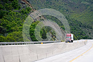 Red big rig semi truck with semi trailer going up hill on winding green highway in mountain