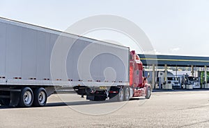 Red big rig semi truck transporting cargo in dry van semi trailer running on the truck stop to fuel station for refuel and photo