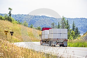 Red big rig semi truck transporting lumber wood on two semi trailers driving on winding road with hills