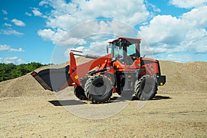 A red-black front-end loader with small wheels against the background of a large pile of stone sand