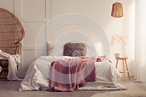 Red blanket on bed with cushions in white bedroom interior with lamp and rattan chair