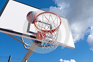 red and blue ball in a basketball hoop with white backboard against a blue sky with cloud