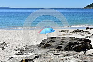 Red and blue beach umbrella on a beach with rocks, bay and blue sky. Cee, Rias Altas, Galicia, Spain.