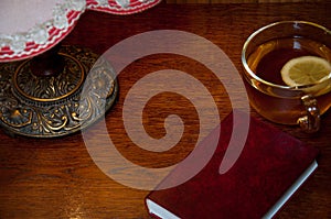 Red book, cup with tea and old vintage lamp on wooden background table at home in the evening. Horizontal view. Space for text.