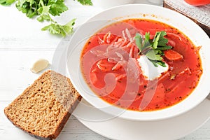 Red borscht with sour cream and herbs in a white plate,  bread on a white  table
