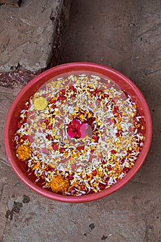 Red bowl with hand washing water on floor, top view. Bright flower petals soaking in water. Indian culture, spa care.