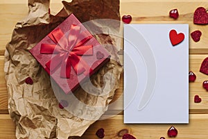 Red box with a gift and next to a white blank sheet for text with a heart on a wooden surface.