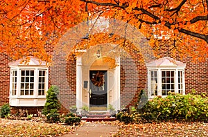 Red brick house entrance with seasonal wreath on door and porch and bay windows on autumn day with leaves on the ground and hydrag