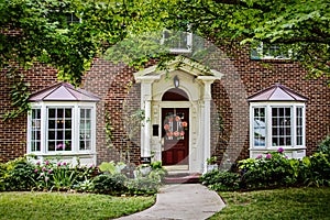 Red brick house with red door and bay windows and pretty flower beds and wreath on door and maple trees in front yard