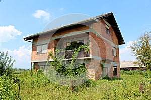 Red bricks abandoned family house with broken windows and front wire fence overgrown with high uncut grass and crawler plants