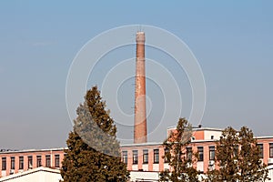 Red bricks tall industrial chimney with metal stairs mounted on side rising high above elongated old factory building