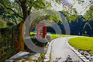 A red British Telephone Booth along a walking path in the park near a brown wooden fence surrounded by lush green trees