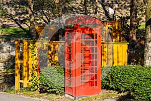 A red British Telephone Booth along a walking path in the park near a brown wooden fence surrounded by lush green trees