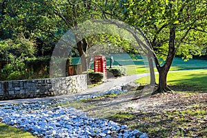 A red British Telephone Booth along a walking path in the park near a brown wooden fence surrounded by lush green trees