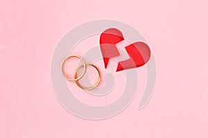 Red broken heart split in two pieces with pair of golden wedding rings on pink background