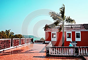 Red building and terrace with orange tiles among tropical vegetation and palm trees by the sea in Goa