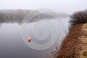 Red buoy in a calm water, trees in mist in the background. River Corrib, Galway city, Ireland. Fall season