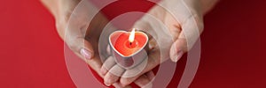 Red burning candle in the shape of a heart in female hands on red background