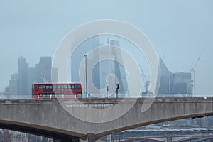 The red bus on the bridge in the foreground and the skyscrapers covered in fog