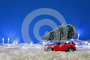 Red car carrying a christmas tree over the snow with garland lights.