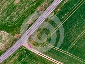 Red car is parking on the green field near the asphalt road. Aerial top down view, agriculture theme. Fresh green color at spring