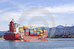 A red cargo ship loaded with multicolored shipping containers enters the port