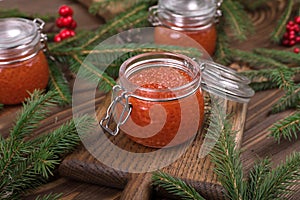 Red caviar in a glass jar on a seasoned white wooden background with a Christmas tree.