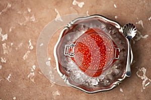 Red caviar in a glass jar with a yoke in a vintage metal dish filled with crushed ice with a curly spoon lying next to