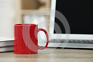 Red ceramic mug, notebooks and laptop on wooden table indoors. Space for text