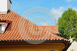 Red ceramic shingles roof with attic windows and chimney against blue sky. Decorative rain gutter.