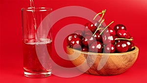 Red cherry juice being poured in a glass and red ripe cherries in wooden bowl on red background