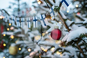 Red Christmas ball decor on a snow-covered Christmas tree with Christmas lights and Finnish flags