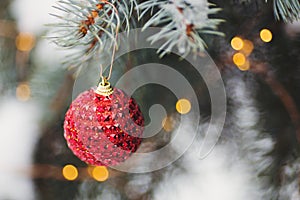 Red Christmas ball hanging on spruce branch covered with snow with warm garland lights