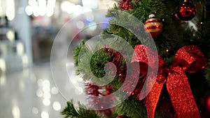 Red Christmas ball with red bow on green Christmas tree close up on blurred shopping Mall background with lights.