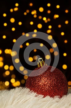 Red Christmas ball with sparkles on fur, defocused lights on back on black background. New Year