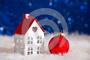 Red Christmas ball with toy little house on white fur with garland lights on blue bokeh background