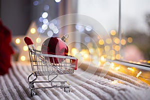 Red Christmas ball in toy supermarket trolley in daylight with warm garland bokeh on background.