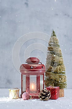 Red Christmas lantern glows beside a festive tree, ornaments, pinecone and gifts in a snowy winter setting. Holiday composition