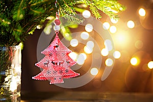 Red Christmas tree toy on a branch of a natural fir tree with lights of garlands in defocus in the background. Metal toy with