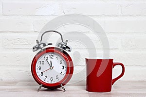 Red classic alarm clock and red coffee mug on white brick wall