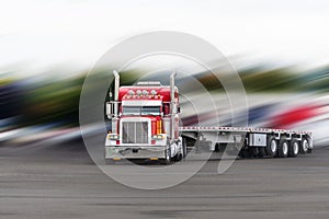 Red classic big rig semi truck with flat bed semi trailer turning on the parking lot on blurry background