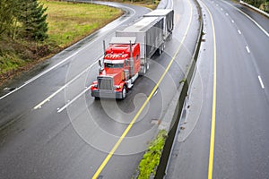 Red classic big rig semi truck with spoiler running on the turning wet road transporting two semi trailers with commercial cargo