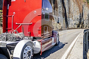 Red classic big rig semi truck tractor with high cab transporting flat bed semi trailer running on the mountain road around rock