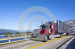 Red classic bonnet big rig semi truck transporting frozen cargo in bright refrigerated semi trailer driving on the truss bridge