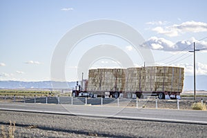 Red classic powerful big rig semi truck transporting pressed hay on two flat bed semi trailer driving on the straight highway road