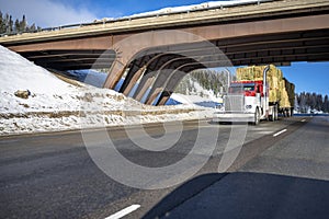 Red classic well maintained big rig semi truck with high chrome pipes transporting hey on the step down semi trailer running under