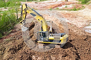 Red clay mining. In a quarry, a yellow excavator with a large bucket loads clay into a truck